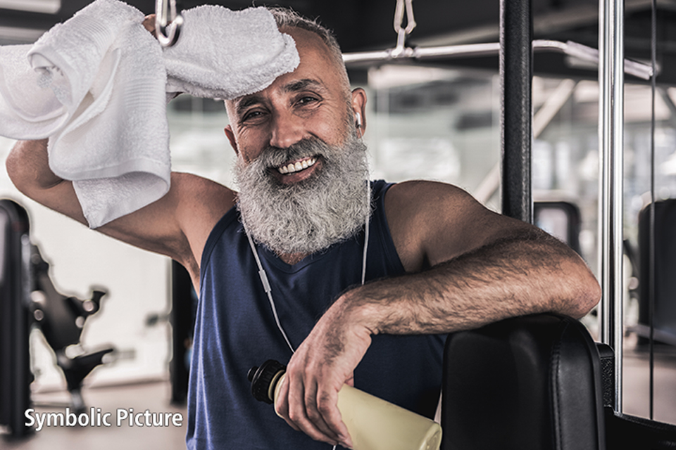 The picture shows an older man with a long, grey beard wiping off his sweat with a towel in the gym after training. He is smiling contentedly at the camera, holding a water bottle and wearing headphones. The background is blurred, but shows other fitness equipment. It is a symbolic image.