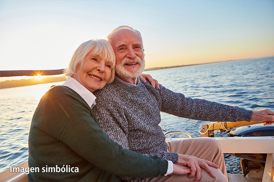 La imagen muestra a una pareja de ancianos abrazados en un barco al atardecer. Parecen felices y contentos, saboreando el momento. El mar de fondo y el cálido sol del atardecer crean una atmósfera romántica y apacible. La imagen transmite un sentimiento de amor, seguridad y tranquilidad en la vejez.